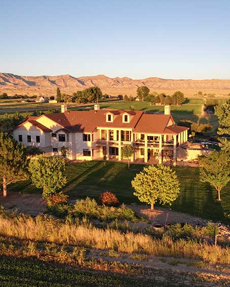 Aerial view of a large house with a red roof, surrounded by green lawns and trees. In the background, there are rolling hills and a clear blue sky, creating a serene landscape.