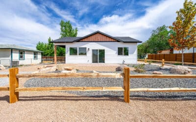 A small white house with black trim and a gable roof sits behind a wooden fence, surrounded by a gravel yard with rocks and minimal landscaping under a blue sky with scattered clouds.