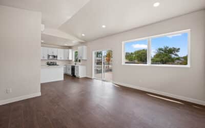 Bright, modern open-concept living area with dark wood floors, white walls, large windows, recessed lighting, and a white kitchen with stainless steel appliances. A sliding glass door opens to an outdoor patio.