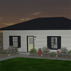 A small, single-story accessory dwelling unit with white siding, black shutters, and a dark roof, surrounded by shrubs and a green lawn, is shown at dusk with a lit porch light.