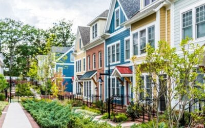 A row of colorful, modern townhouses with red, blue, and yellow exteriors, landscaped front gardens, small porches, and a sidewalk lined with greenery and trees on a bright, cloudy day.